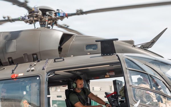 A military helicopter, consistent with a UH-72 Lakota or similar utility rotary-wing aircraft, is shown on the ground with its side door open. A crew member in military flight gear and aviation headset is seated in the cabin, while at least one additional personnel is visible in the cockpit area. The aircraft is painted in standard military olive/gray livery.