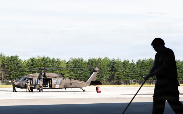 A silhouetted figure sweeps a tarmac surface in the foreground of a military airfield. In the background, a military helicopter — consistent with a UH-60 Black Hawk — is parked on the flight line with personnel nearby. The scene is set against a treeline under an overcast sky.