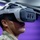 A person wearing military camouflage (MultiCam/OCP pattern) uniform is using an advanced VR/XR headset, likely in a training or demonstration environment. The headset appears to be a modified or military-grade variant with multiple forward-facing cameras and sensors mounted on the front. The purple backdrop suggests a controlled indoor facility, possibly a military training or technology demonstration space.