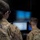 Two military personnel in OCP (Operational Camouflage Pattern) uniforms are working at a computer workstation with dual monitors. One individual is wearing headphones and operating a laptop, while the other observes from behind. The screens appear intentionally blurred, likely for operational security purposes.