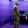 A military personnel wearing camouflage uniform and tan boots is using a virtual reality (VR) headset in a purple-lit enclosed space. The setup includes a desk with two monitors and a laptop, suggesting a military VR training or simulation environment. A cable from the VR headset trails along the floor connecting to the workstation.