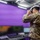 A military personnel member wearing camouflage uniform is using a VR (virtual reality) headset in what appears to be a dedicated VR training or simulation room with a bright purple background. The room is equipped with computer monitors and a desk setup, suggesting this is a technology-enabled training environment. A cable runs from the VR headset, indicating a tethered, high-performance VR system.