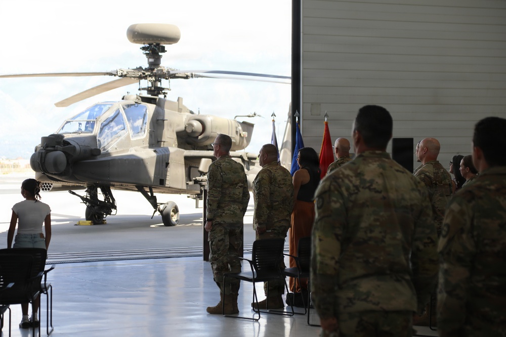 Military ceremony held in aircraft hangar with AH-64 Apache helicopter as backdrop