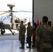 Military ceremony held in aircraft hangar with AH-64 Apache helicopter as backdrop