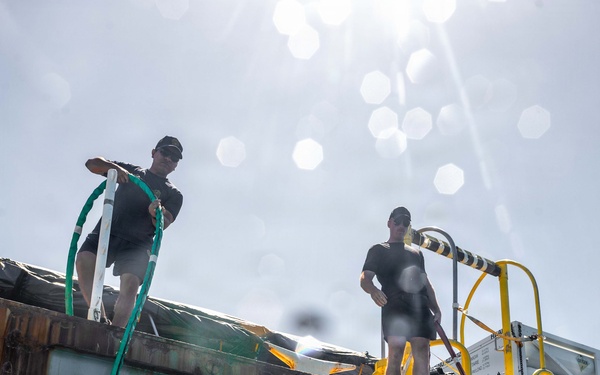 A commercial or military diver in a full diving helmet and dry suit is entering the water from a rusted barge or work platform. Two surface support personnel dressed in black shirts and caps stand on the deck above, one managing a green umbilical/hose line. The photo is taken from a split water-level perspective, capturing the dramatic water splash of the diver's entry.