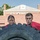 Two young people wearing red shirts are pushing or flipping a large tractor tire outdoors, appearing to be engaged in a fitness or training exercise. Both individuals display focused, determined expressions as they lean into the heavy rubber tire. The setting appears to be an outdoor area near a brick building.