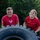 Two young people wearing matching red t-shirts are working together to flip or push a large Michelin tractor/agricultural tire outdoors. The male on the left is straining with visible effort while the female on the right appears more composed. The activity appears to be a fitness or team-building exercise taking place on a grassy area with trees in the background.