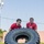 Two individuals wearing matching red t-shirts are performing a tire flip exercise outdoors on a grassy area. They are pushing a large, heavy tractor-style tire in what appears to be a fitness training or physical conditioning session. Both individuals display visible physical exertion on their faces.