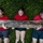 Three young people wearing matching red t-shirts are lying on a grass field covered in clover, collectively holding up a large, weathered wooden log above their bodies. They appear to be engaged in a team physical challenge or training exercise, showing visible strain and effort. Their clothing and skin show signs of dirt and mud, suggesting prior physical activity.