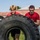 Two young men wearing red t-shirts are collaboratively flipping a large heavy-duty tractor tire on a grassy outdoor surface. A third person in a black t-shirt observes from the background. The activity appears to be a strength and conditioning exercise or fitness training drill.