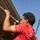 A young man wearing a red t-shirt with partial text reading 'MAR...' and 'THE FEW...' is climbing or pulling himself up a thick rope outdoors. The photo is taken from a low angle looking upward, emphasizing his physical effort and upper body strength. The setting appears to be near a brick building under a clear blue sky.