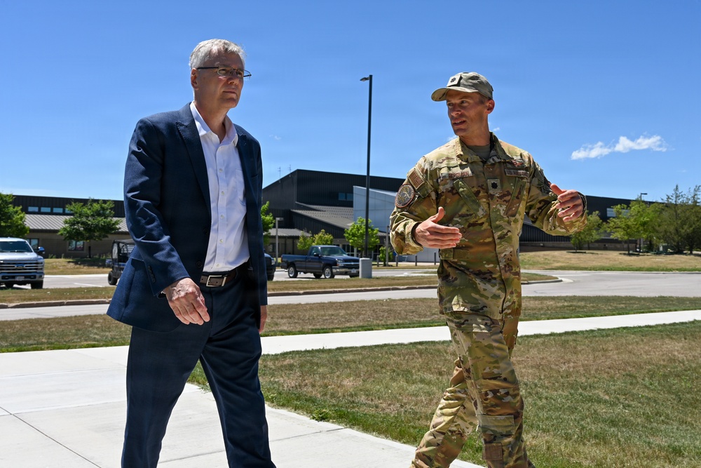 Two individuals are walking outdoors on a sidewalk at what appears to be a military installation or government facility. A civilian in a navy blue suit is accompanied by a military personnel wearing OCP (Operational Camouflage Pattern) uniform, who appears to be briefing or explaining something. The setting suggests an official visit or tour of the facility.