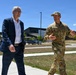 Two individuals are walking outdoors on a sidewalk at what appears to be a military installation or government facility. A civilian in a navy blue suit is accompanied by a military personnel wearing OCP (Operational Camouflage Pattern) uniform, who appears to be briefing or explaining something. The setting suggests an official visit or tour of the facility.
