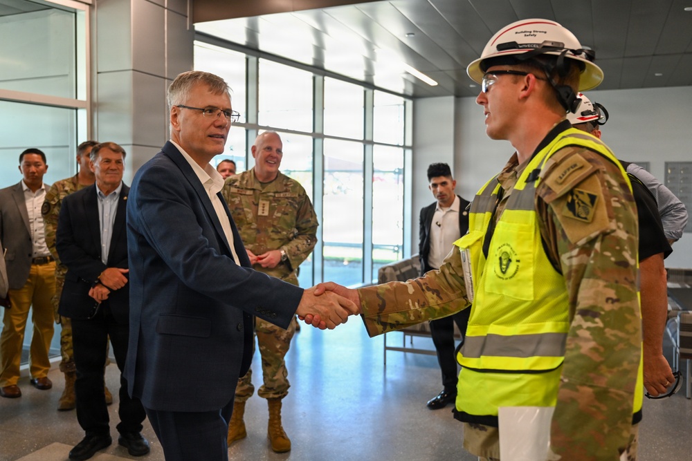 A civilian in a navy blue suit is shaking hands with a military soldier wearing camouflage uniform, a high-visibility yellow safety vest, and a white hard hat. The setting appears to be inside a modern building, likely a facility under construction or recently completed. Several other military personnel and civilians are visible in the background observing the exchange.