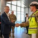 A civilian in a navy blue suit is shaking hands with a military soldier wearing camouflage uniform, a high-visibility yellow safety vest, and a white hard hat. The setting appears to be inside a modern building, likely a facility under construction or recently completed. Several other military personnel and civilians are visible in the background observing the exchange.