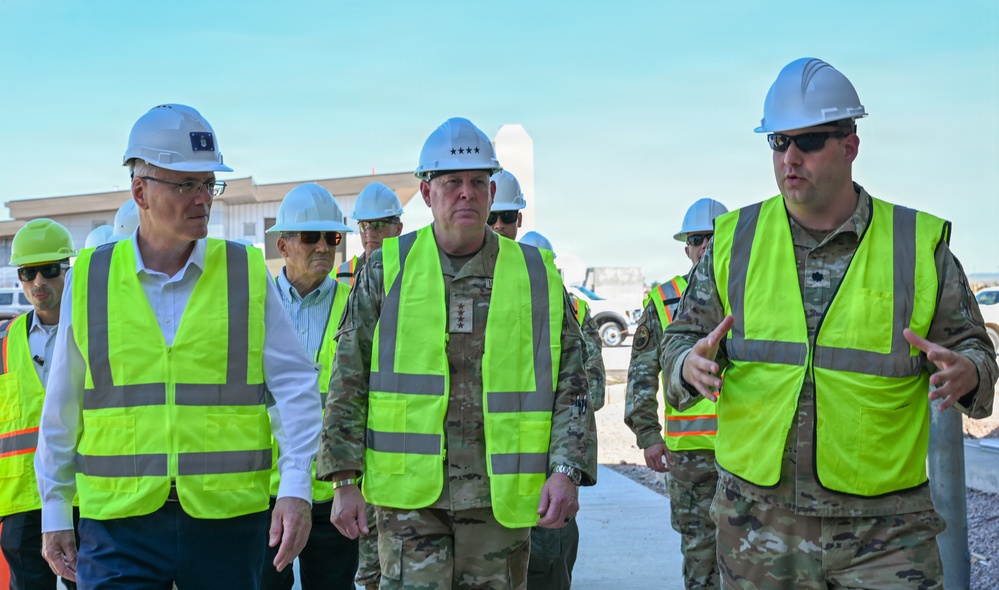 A group of individuals wearing white hard hats and high-visibility yellow safety vests are conducting a site walkthrough at what appears to be a military construction or infrastructure project. The group includes uniformed military personnel, with at least one individual displaying four-star rank insignia on their uniform, alongside civilians. A person on the right appears to be briefing or guiding the group.