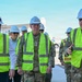 A group of individuals wearing white hard hats and high-visibility yellow safety vests are conducting a site walkthrough at what appears to be a military construction or infrastructure project. The group includes uniformed military personnel, with at least one individual displaying four-star rank insignia on their uniform, alongside civilians. A person on the right appears to be briefing or guiding the group.
