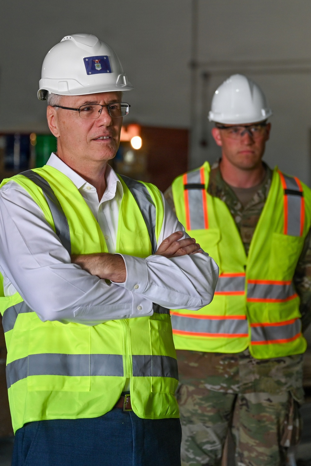 Two individuals are standing inside what appears to be a military facility or construction site. The foreground figure wears civilian attire with a white hard hat bearing a U.S. Air Force insignia sticker, while the background figure is in military camouflage uniform. Both are wearing high-visibility safety vests, suggesting an official site inspection or tour.