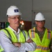 Two individuals are standing inside what appears to be a military facility or construction site. The foreground figure wears civilian attire with a white hard hat bearing a U.S. Air Force insignia sticker, while the background figure is in military camouflage uniform. Both are wearing high-visibility safety vests, suggesting an official site inspection or tour.