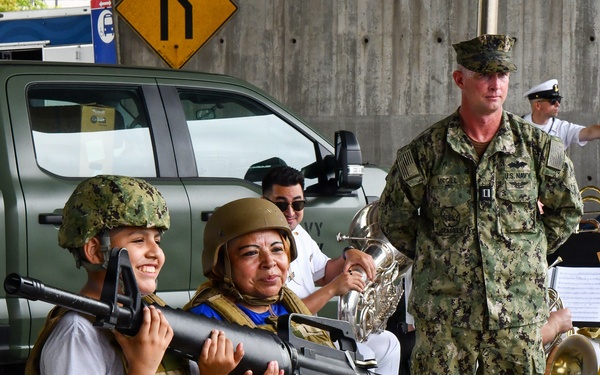 A public military outreach or open house event where civilians, including a child and an adult woman, are trying on tactical gear (helmets and plate carriers) and posing with what appears to be a training/display rifle. U.S. Navy personnel are present, including a Seabee officer in NWU Type III camouflage and white-uniformed band members with brass instruments in the background. The setting appears to be an outdoor covered area, likely a military base or public event venue.