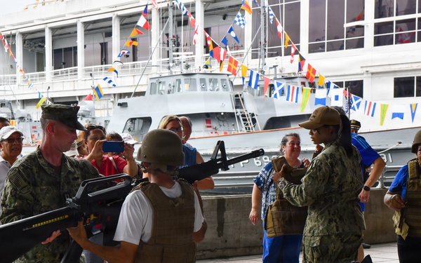 A military open-day or public outreach event at a naval dock where uniformed personnel are demonstrating equipment to civilians. A Navy servicemember is showing a rifle to a child wearing a tactical vest and helmet, while another servicemember assists a civilian woman in trying on body armor. Naval vessels decorated with signal flags are visible in the background.