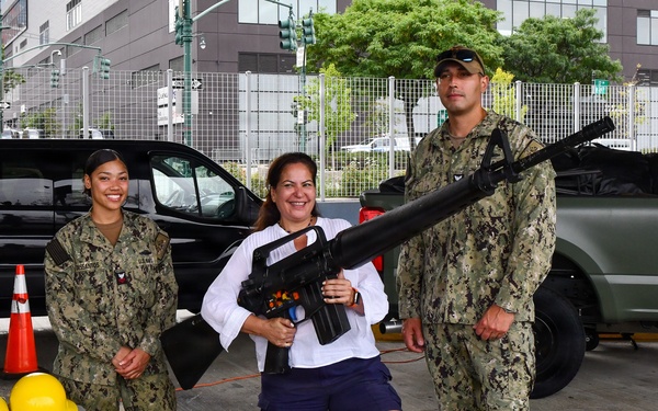 Three individuals pose for a photo in an outdoor urban setting, likely during a public military outreach or recruitment event. Two personnel in U.S. Navy NWU Type III (digital camouflage) uniforms flank a civilian woman who is holding what appears to be a large prop or training replica firearm. A folding table with safety equipment is visible to the left.