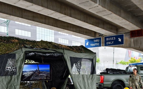 Two military personnel in Navy working uniforms are setting up or manning a tactical display station under an elevated highway or overpass structure. The setup includes a camouflage-netted tent shelter attached to a military green pickup truck, with equipment and monitors visible. The location appears to be an urban area, likely during a public event such as Fleet Week or a military demonstration.