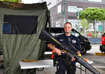 A police officer in dark blue uniform is posing outdoors at what appears to be a public safety or law enforcement exhibition, holding a large rifle with an attached grenade launcher. The setting includes a camouflage-covered tent, folding tables, and military-style vehicles in the background. The event appears to be taking place in an urban area with commercial buildings visible.