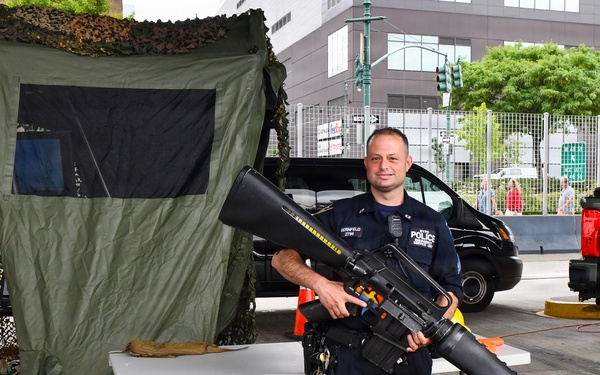 A police officer in dark blue uniform is posing outdoors at what appears to be a public safety or law enforcement exhibition, holding a large rifle with an attached grenade launcher. The setting includes a camouflage-covered tent, folding tables, and military-style vehicles in the background. The event appears to be taking place in an urban area with commercial buildings visible.