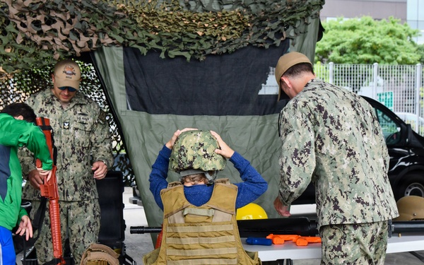 A military public outreach or recruitment event where U.S. Navy personnel are allowing children to try on military gear including tactical vests and helmets. Two sailors in Navy Working Uniform (NWU Type III camouflage) are assisting young visitors under a camouflage netting canopy. A display table with various military equipment is visible in the background.