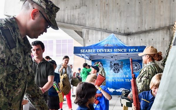 A military public outreach event featuring U.S. Navy Seabees interacting with children and civilians. A booth for the 'Underwater Construction Team' (Seabee Divers Since 1974) is visible in the background. Children are handling toy/replica weapons and equipment as part of an educational display.