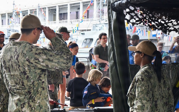 A public military open house or fleet week event at a waterfront dock, where U.S. Navy personnel in NWU Type III camouflage uniforms interact with civilian visitors including children. Two sailors are saluting while children, including one in a Cub Scout uniform, examine equipment on a display table. Naval vessels and international signal flags are visible in the background.