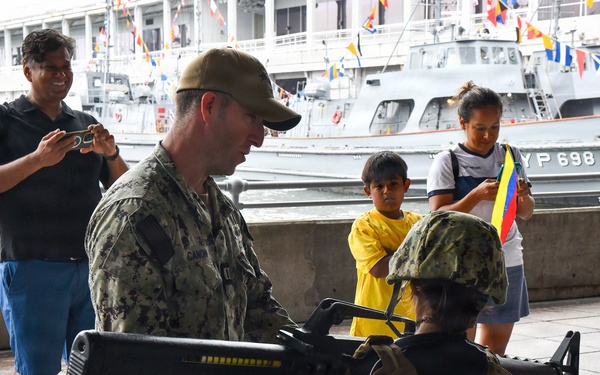 A military or naval open-day event at a harbor/port facility where personnel in camouflage uniforms are demonstrating equipment to civilians. Two service members are handling what appears to be an M16-style rifle, while civilians including a child and adults observe and photograph the display. Naval vessels decorated with signal flags are visible in the background, including one marked 'YP 698'.