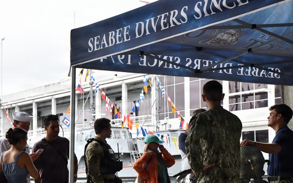 A public outreach or open house event featuring a 'Seabee Divers' display booth at a waterfront naval facility. Military personnel in camouflage uniforms are engaging with civilian visitors, including families with children, while showcasing diving equipment. Naval vessels decorated with signal flags are visible in the background.