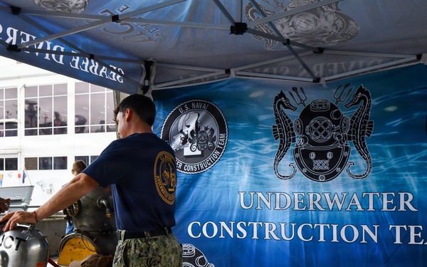 A U.S. Navy Underwater Construction Teams (UCT) display booth set up under a canopy tent, likely at a public outreach or fleet week event. A Navy sailor in camouflage utilities is showing diving equipment to a civilian visitor at a table. The backdrop features a large blue banner with UCT insignia, logos, and the text 'Underwater Construction Teams.'