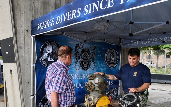 A U.S. Navy Seabee Diver is presenting diving equipment at an outdoor display booth labeled 'Seabee Divers Since 19[47]' to a civilian visitor. The booth features a backdrop with U.S. Naval Underwater Construction Team insignia and Pacific Fleet logos. Two vintage and modern diving helmets are displayed on a folding table along with other diving-related equipment.