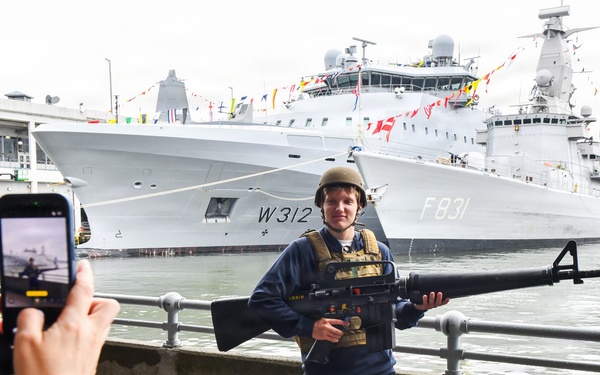 A young civilian man poses for a photo at what appears to be a naval open day or fleet review event. He is wearing a military helmet and tactical vest while holding a large rifle, likely props provided for tourist photos. Two naval vessels are visible docked in the background, one marked 'W312' and another marked 'F831', decorated with signal flags.