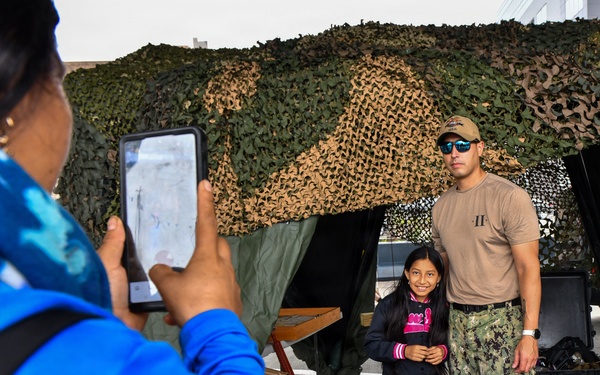 A military service member in U.S. Navy camouflage uniform and tan t-shirt poses for a photo with a young girl in front of a camouflage netting backdrop. A person in a blue jacket is photographing them using a tablet device. The setting appears to be a military open house, public outreach event, or ship tour.
