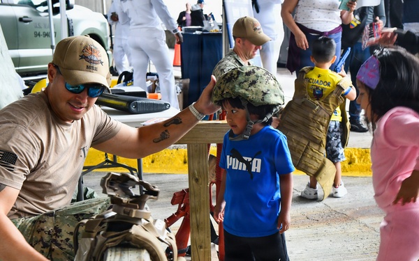 A U.S. Navy service member crouches down and places a military helmet on a young boy's head during what appears to be a public outreach or community engagement event. Children and adults are gathered in an outdoor area near military equipment displays, with one child in the background wearing a tactical vest labeled 'Ecuador'. The event appears to be a military open house or joint exercise demonstration.