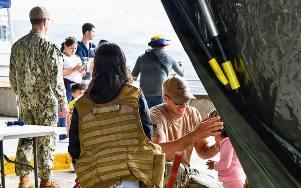 A military open house or community outreach event at what appears to be a naval facility or pier. Personnel in U.S. Navy digital camouflage uniforms interact with civilian visitors, including families with young children. A display table in the foreground features miniature naval ship models and toy items.