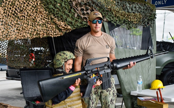 A military service member in U.S. Navy digital camouflage uniform assists a young child in holding a rifle at what appears to be a public military outreach or recruitment event. The child is wearing a military helmet and tactical vest, suggesting a hands-on demonstration setup for civilian visitors. The setting includes camouflage netting, military vehicles, and display tables with equipment.