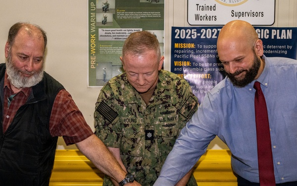Three men are cutting a birthday cake at what appears to be a U.S. Navy facility. A Navy officer in camouflage uniform, identified by name tape as 'YOUNG,' is cutting the cake alongside a civilian in a dress shirt and tie, while another civilian with a beard observes. A ceremonial sword is being used to cut the cake, which reads 'Happy 40th Birthday' with what appears to be a Navy emblem.