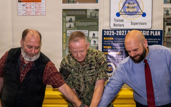 Three individuals are cutting a decorated birthday cake at a table covered with a blue tablecloth in what appears to be a military facility. A U.S. Navy officer in camouflage uniform, identified by name tape as 'YOUNG,' is flanked by two civilians participating in the cake-cutting ceremony. A ceremonial sword is visible on the table, and background posters reference a '2025-2027 Strategic Plan' related to Pacific Fleet SSBNs.