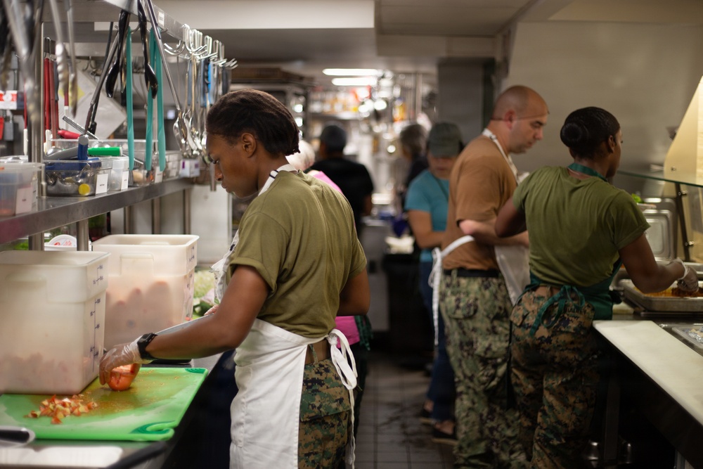 A busy military kitchen with several personnel wearing camouflage uniforms and white aprons engaged in food preparation. The foreground shows a service member chopping tomatoes on a green cutting board, while others work at various stations in the background. The setting appears to be a military mess hall or galley kitchen with stainless steel shelving and equipment.