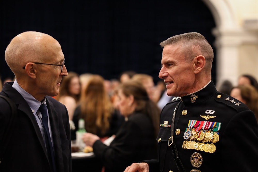 Two men are engaged in conversation at what appears to be a formal event or reception. One man is in civilian attire while the other is wearing a U.S. Marine Corps dress blue uniform adorned with numerous medals, ribbons, and rank insignia indicating a high-ranking officer. The background shows a crowded indoor venue with many attendees.