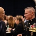 Two men are engaged in conversation at what appears to be a formal event or reception. One man is in civilian attire while the other is wearing a U.S. Marine Corps dress blue uniform adorned with numerous medals, ribbons, and rank insignia indicating a high-ranking officer. The background shows a crowded indoor venue with many attendees.