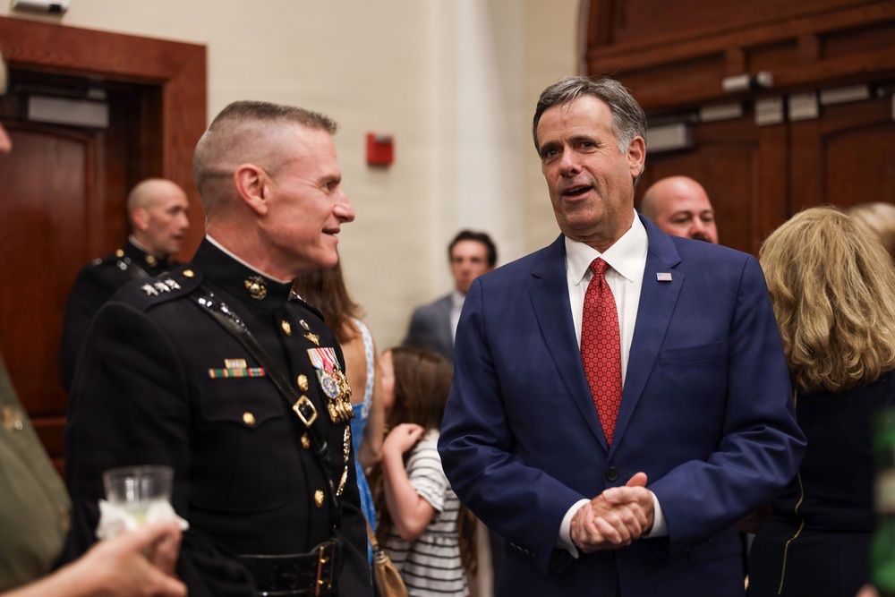 Two men are engaged in conversation at what appears to be a formal indoor reception or event. One man is wearing a U.S. Marine Corps dress uniform with multiple medals and ribbons, while the other is dressed in a navy blue suit with a red tie and an American flag lapel pin. The setting appears to be a formal government or institutional building with wood-paneled walls.