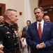 Two men are engaged in conversation at what appears to be a formal indoor reception or event. One man is wearing a U.S. Marine Corps dress uniform with multiple medals and ribbons, while the other is dressed in a navy blue suit with a red tie and an American flag lapel pin. The setting appears to be a formal government or institutional building with wood-paneled walls.