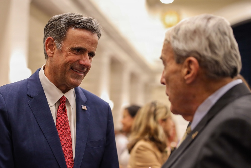 Two older men are engaged in a conversation in what appears to be a formal indoor setting, likely a conference hall or government building. The man on the left is smiling and wearing a navy blue suit with a red patterned tie and a small American flag lapel pin. The background shows a blurred crowd of people in a well-lit corridor.