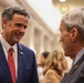 Two older men are engaged in a conversation in what appears to be a formal indoor setting, likely a conference hall or government building. The man on the left is smiling and wearing a navy blue suit with a red patterned tie and a small American flag lapel pin. The background shows a blurred crowd of people in a well-lit corridor.